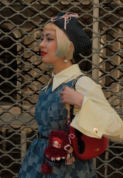 Una persona con un vestido azul decorado, con un cuello blanco y mangas amarillas, lleva una bolsa roja con un llavero de muñeca, y lleva un sombrero de rayas oscuras.