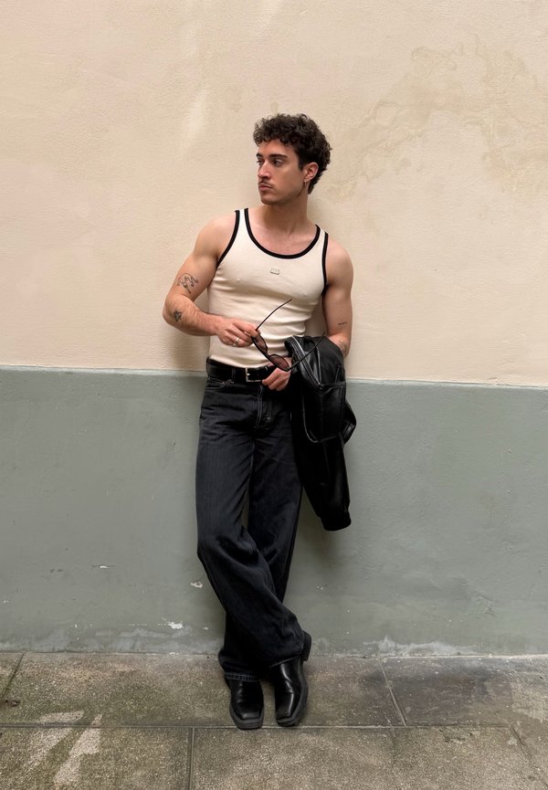 Νεαρός με σγουρά μαλλιά ακουμπά σε διχρωμικό τοίχο, φορώντας λευκή φανέλα, μαύρο παντελόνι, κρατώντας γυαλιά ηλίου και μαύρο σακάκι.