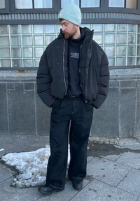 Veste matelassée noire, sweat-shirt foncé, jean noir ample et bottes noires. Bonnet bleu clair. Neige au sol, mur en briques de verre en arrière-plan.