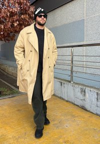 Beige, dupla gombos trench kabát, fekete pulóver, sötétzöld nadrág és fekete csizma; napszemüveg és fekete sapka fehér mintával.