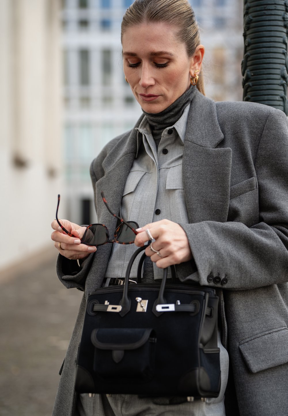 Svart handväska med silverdetaljer, mjuk textur och strukturerad form. Grå ullrock över en ljusgrå skjorta med knappar och polokrage.