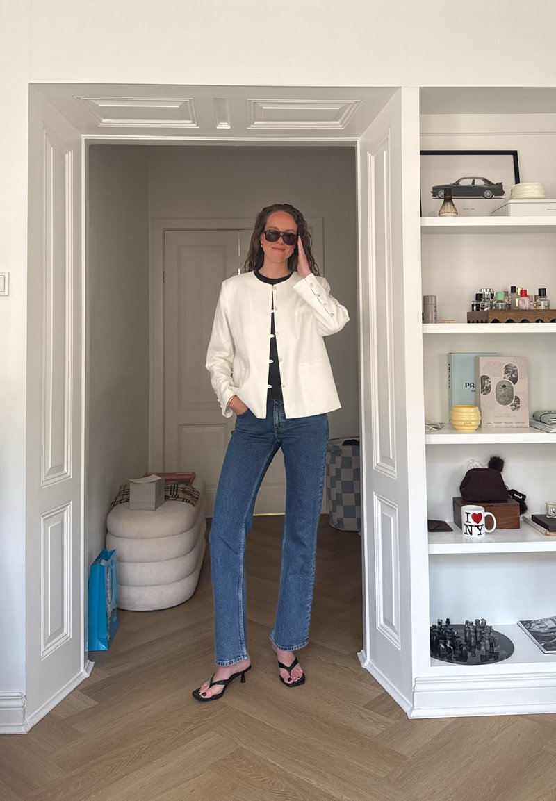 Femme portant des lunettes de soleil, une veste blanche, un haut noir, un jean bleu et des sandales noires se tenant dans une embrasure de porte, avec des étagères et de la décoration à droite.