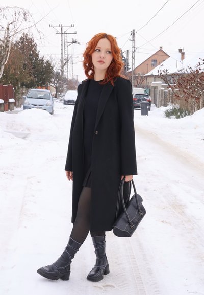 Mujer pelirroja con abrigo negro, vestido y botas, sosteniendo un bolso, de pie en una calle residencial nevada con coches estacionados y casas.