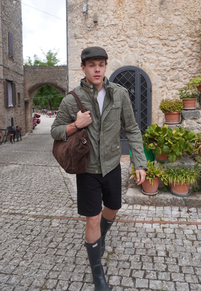 Joven con chaqueta verde, pantalones cortos negros, botas de goma y gorra camina por una calle adoquinada llevando una bolsa marrón sobre el hombro cerca de edificios de piedra.