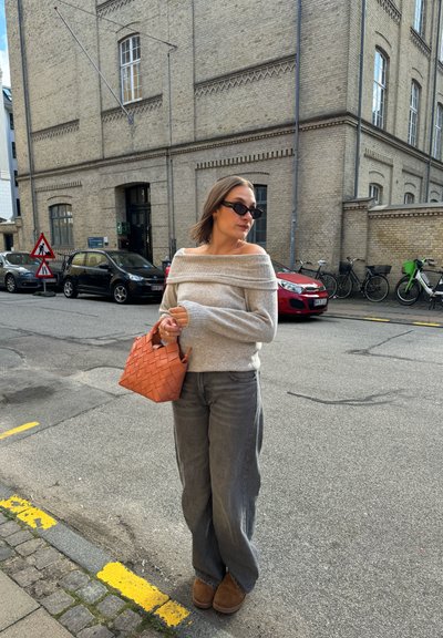 Suéter gris claro sin mangas, jeans gris de pierna ancha, botas marrones hasta el tobillo y un bolso tejido naranja. Entorno urbano en la calle con coches.