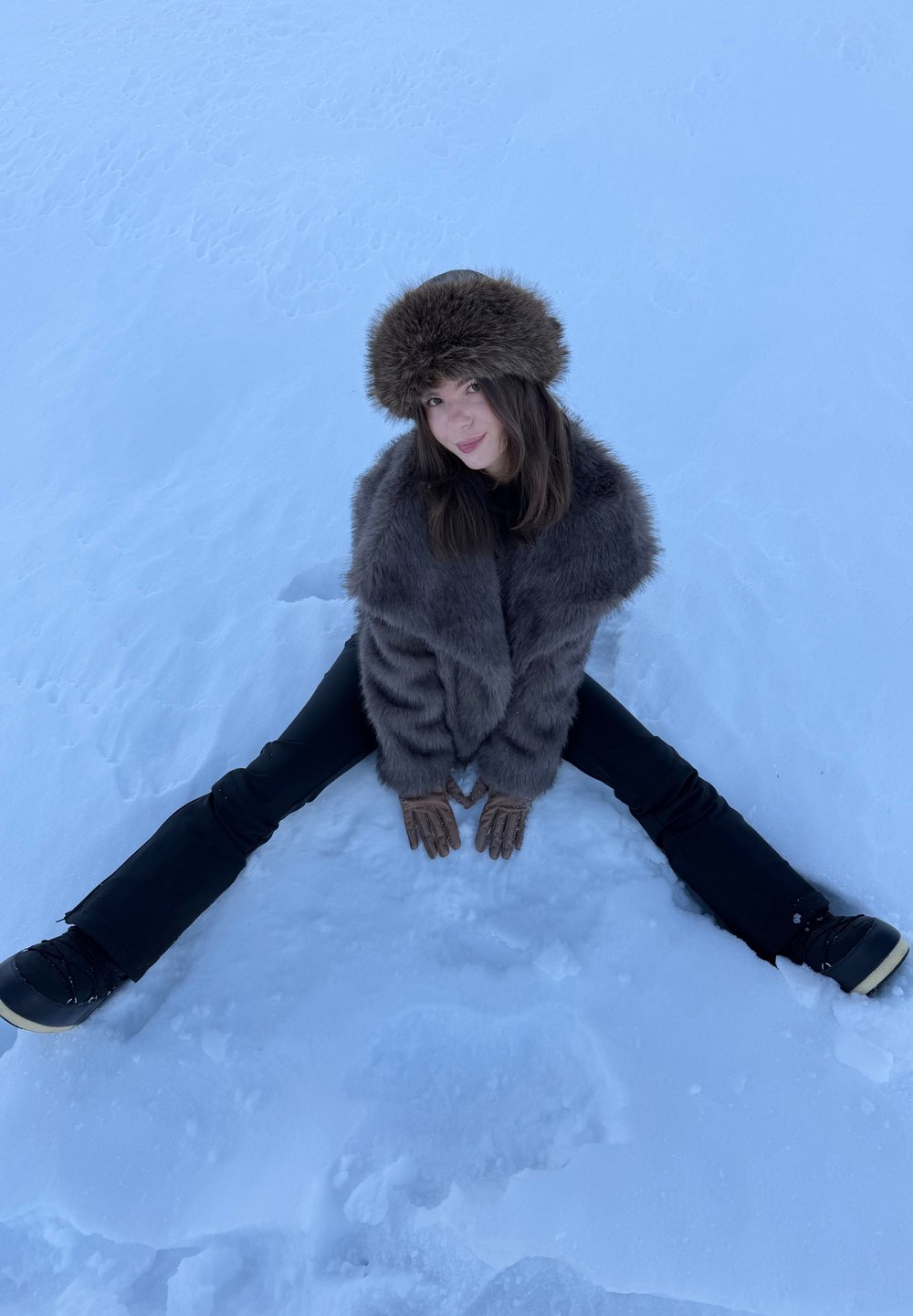 Jeune femme portant un chapeau en fourrure et un manteau, avec des gants et des bottes de neige, assise les jambes écartées sur un sol enneigé.