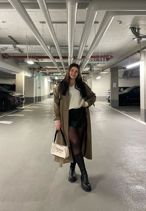 Lang beige trenchcoat over en hvid sweater, sort mini nederdel og knæhøje støvler; holder en cremet håndtaske med "GUESS" logo.