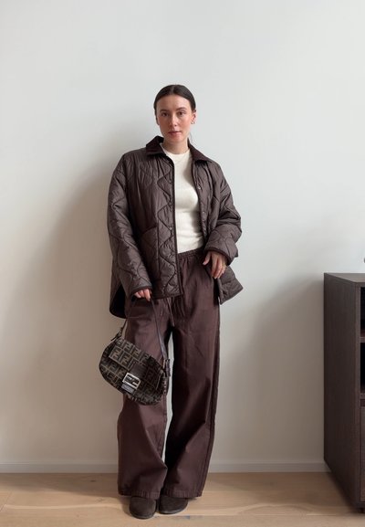 Mujer de pie contra una pared lisa, con chaqueta acolchada marrón, pantalones de pierna ancha, top blanco, sosteniendo un bolso estampado a un lado.