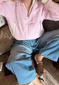 Veste zippée rose côtelée avec accents blancs, portée sur un pantalon large bleu et des chaussures slip-on marron doublées de fourrure, sur un canapé en velours.