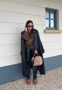 Μαύρο δερμάτινο παλτό τρεντς πάνω από ένα καφέ πουλόβερ με μοτίβο, μαύρο παντελόνι από ψεύτικο δέρμα, καφέ σουέτ μπότες και μια υφασμάτινη μπεζ τσάντα.