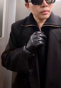 Fekete kabát bordázott gallérral, cipzáras elülső résszel; fekete bőrkesztyűt visel, ezüst liftben áll. Simább textúra, minimalista dizájn.