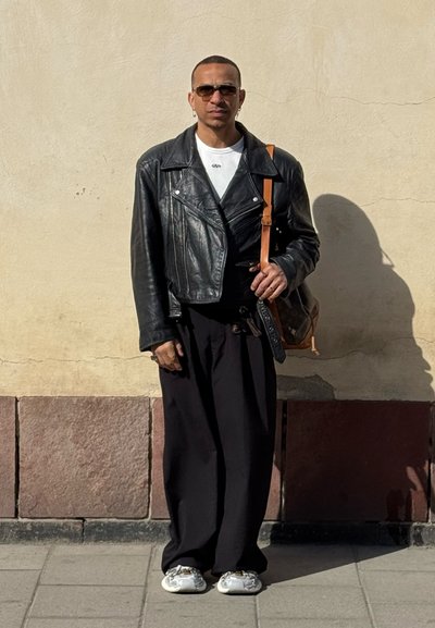 Persona con chaqueta de cuero negra de gran tamaño, camisa blanca, pantalones negros holgados, zapatillas blancas, gafas de sol y llevando un bolso marrón al hombro, de pie afuera.
