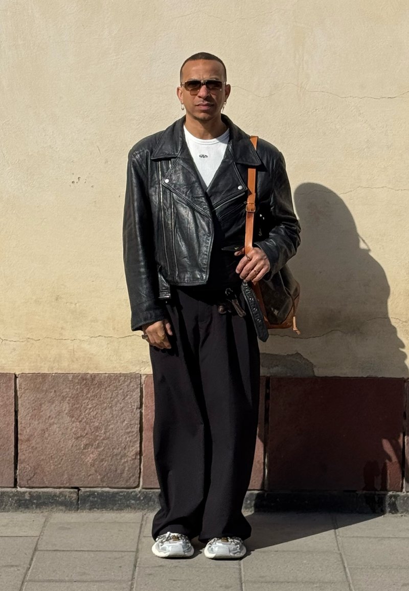 Personne portant une veste en cuir noire oversize, une chemise blanche, un pantalon noir ample, des baskets blanches, des lunettes de soleil et portant un sac à bandoulière marron, debout à l'extérieur.