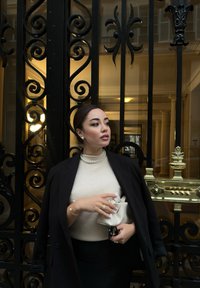 Cappotto nero sopra un maglione beige, gonna nera; tiene una borsa crema. Perle accentuano collo e orecchie. Dietro una complessa cancellata in ferro battuto nera.