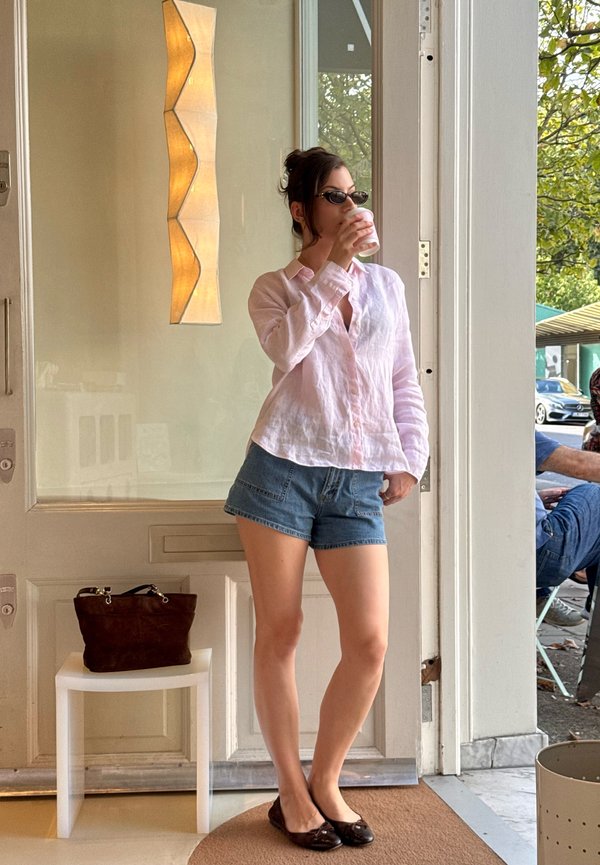 Femme portant des lunettes de soleil, un chemisier rose clair et un short en jean boit dans une tasse, debout près d'une porte avec un sac marron sur un tabouret blanc.