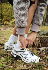 Des chaussures de sport blanches avec des accents noirs et bleus, en matériau léger, à bout arrondi et système de laçage rapide, portées dans un cadre forestier.