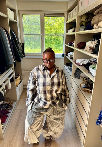 Persona con gafas y camisa de cuadros está en un vestidor con estantes y ropa colgada a ambos lados, luz natural desde la ventana detrás.