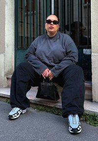 Grå langermet skjorte, oversized svarte bukser, og hvite/grå joggesko. Holder en liten svart håndveske. Minimalistisk urban setting med en grønn dør.