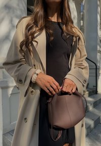 Μπεζ παλτό πάνω από ένα μαύρο φόρεμα, κρατώντας μια μικρή, υφασμάτινη καφέ δερμάτινη τσάντα. Τα μαλλιά είναι μακριά και κυματιστά, με το φως να πέφτει από το πλάι.