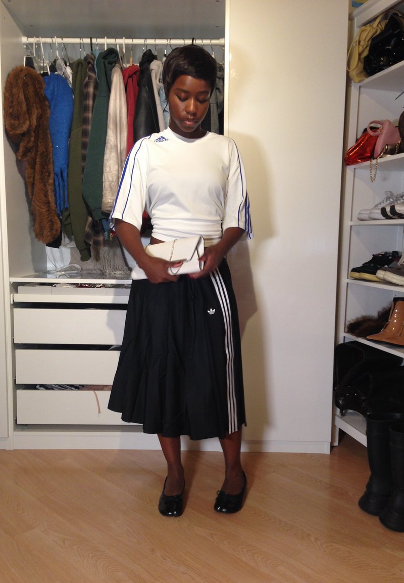 T-shirt de sport blanc avec des rayures bleues, jupe plissée noire avec des rayures blanches sur les côtés, et une pochette blanche simple à la main. Fond en parquet.