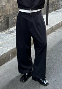 Černé široké kalhoty vyrobené z hladkého materiálu s sklady v přední části, spárované s lesklými černými botami, stojící na dlažební ulici.