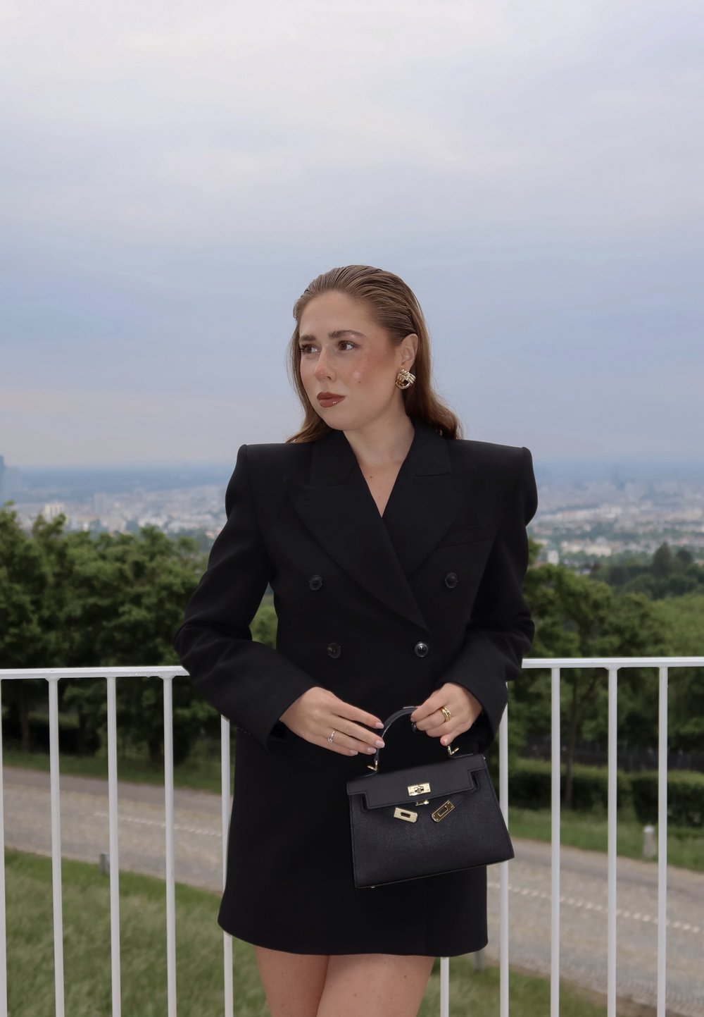 Blazer noir ajusté à double boutonnage, longueur mini, épaules structurées. Tient un petit sac à main noir avec des accessoires et des éléments dorés.