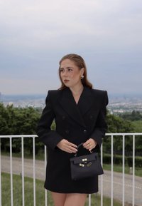 Blazer noir ajusté à double boutonnage, longueur mini, épaules structurées. Tient un petit sac à main noir avec des accessoires et des éléments dorés.