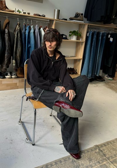 Sudadera negra con cremallera, vaqueros negros desgastados y botas burdeos brillantes. Cadenas y tatuajes visibles. Silla de madera y perchero en el fondo.