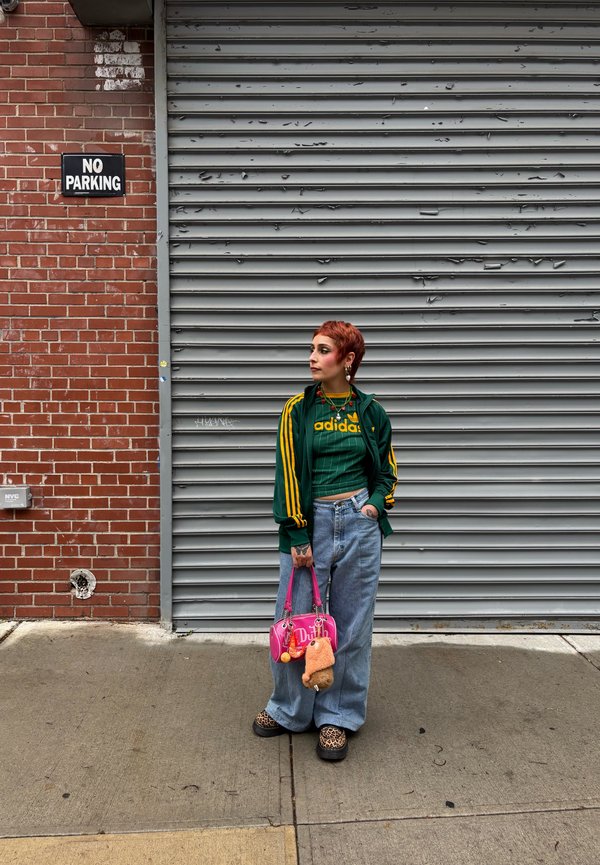 Casaco atlético verde com riscas amarelas, camisa gráfica verde, jeans azuis largos, bolsa rosa e calçado preto com padrão de leopardo.