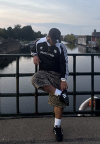 Musta Adidas-takki valkoisilla raidoilla, leopardikuosiset shortsit, mustat sneakersit, joissa on kolme valkoista raitaa, ja asusteita. Ulkoilma-urban ympäristö.