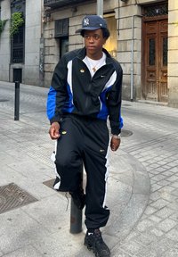 Crni i plavi Nike trenerka s bijelim detaljima, uparena sa sivom baseball kapom New York Yankees i crnim tenisicama. Stoji na stupu.