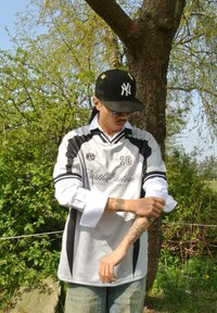 Weißes Fußballtrikot mit schwarzen Streifen, kurzen Ärmeln und dem Logo "North Eastern." Helle blaue Jeans. Schwarze Kappe mit Logo. Sichtbare Armtattoos.