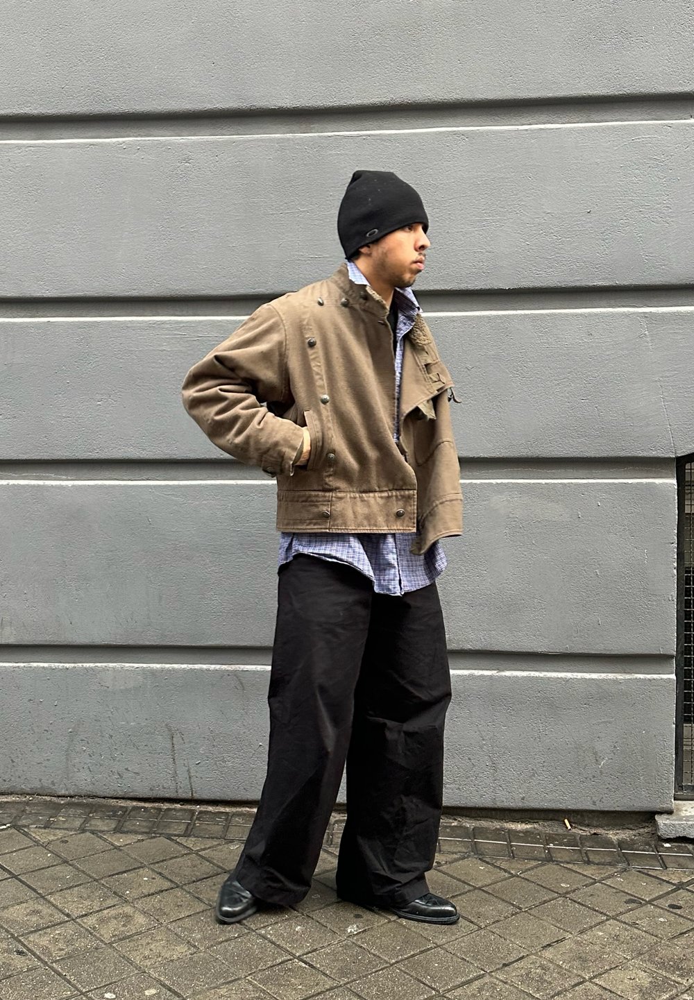 Barna rövidített dzseki gombos részletekkel, alatta kék kockás ing, fekete laza nadrág és fekete sapka, szürke fal előtt.
