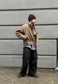 Barna rövidített dzseki gombos részletekkel, alatta kék kockás ing, fekete laza nadrág és fekete sapka, szürke fal előtt.