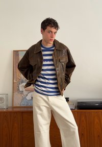 Rjava usnjena jakna z ovratnikom, modro-bela črtasta srajca in bele široke hlače, na leseni podlagi in ozadju z umetniškim motivom.