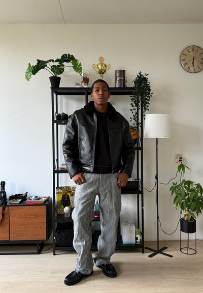 Chaqueta de cuero negro con cuello de borrego, pantalones grises a rayas, mocasines negros. El fondo muestra una estantería con plantas y objetos decorativos.