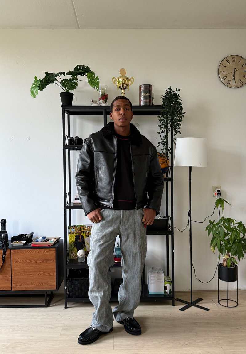 Veste en cuir noire avec col en peau de mouton, pantalon gris à rayures, mocassins noirs. En arrière-plan, une étagère avec des plantes et des objets décoratifs.