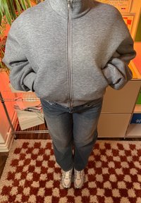 Siva jopica z zadrgo in visokim ovratnikom, kombinirana z ohlapnimi modrimi kavbojkami in sivimi supergami, stoji na vzorčasti preprogi.