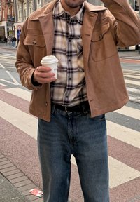 Béžová semišová bunda se dvěma předními kapsami, pod ní kostkovaná košile a modré džíny. Drží bílý jednorázový kelímek na kávu. Městské prostředí.