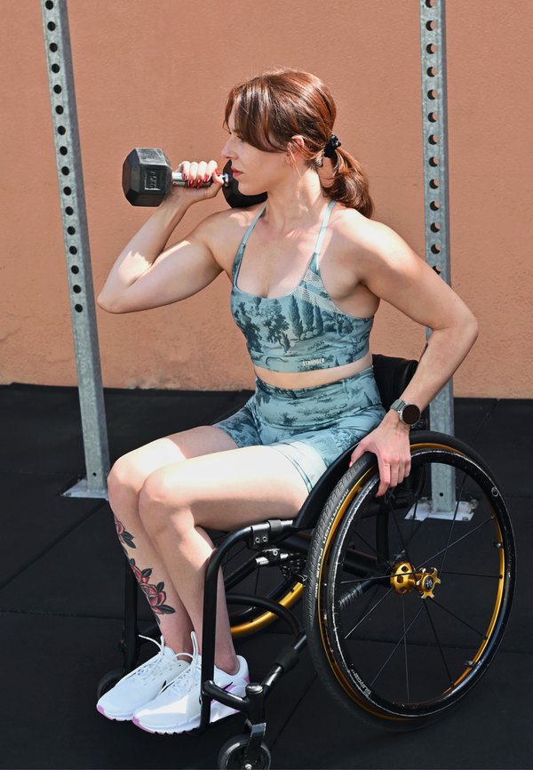 Femme en tenue d'entraînement soulève un haltère assise dans un fauteuil roulant dans un gymnase extérieur avec du matériel d'exercice.