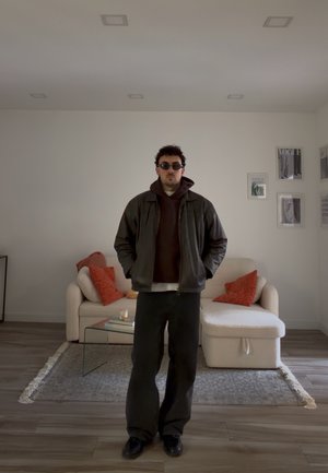 Hombre con gafas de sol, chaqueta oscura y sudadera con capucha, de pie en una sala de estar moderna con sofá beige, cojines naranjas, mesa de centro de cristal y arte enmarcado.