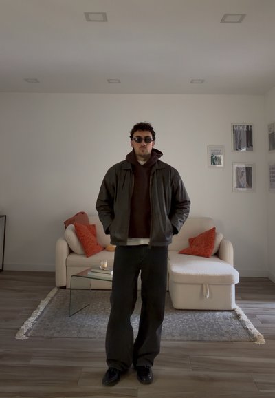 Hombre con gafas de sol, chaqueta oscura y sudadera con capucha, de pie en una sala de estar moderna con sofá beige, cojines naranjas, mesa de centro de cristal y arte enmarcado.