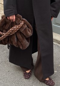Sac en fausse fourrure marron avec poignée tressée, manteau long noir, pantalon large marron foncé, et chaussures violet foncé ornées de clous dorés.