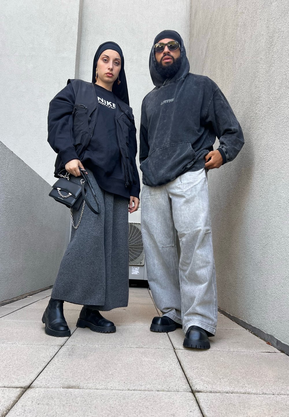Δύο άτομα στέκονται δίπλα-δίπλα. Η γυναίκα φοράει μια μαύρη υπερμεγέθη ζακέτα και γκρι φαρδιά παντελόνια, κρατώντας μια μικρή μαύρη τσάντα. Ο άνδρας φοράει ένα σκούρο hoodie και ανοιχτό γκρι παντελόνια, και τα δύο σε χαλαρή γραμμή. Φοράνε χοντρές μαύρες μπότες.