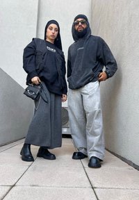 Δύο άτομα στέκονται δίπλα-δίπλα. Η γυναίκα φοράει μια μαύρη υπερμεγέθη ζακέτα και γκρι φαρδιά παντελόνια, κρατώντας μια μικρή μαύρη τσάντα. Ο άνδρας φοράει ένα σκούρο hoodie και ανοιχτό γκρι παντελόνια, και τα δύο σε χαλαρή γραμμή. Φοράνε χοντρές μαύρες μπότες.