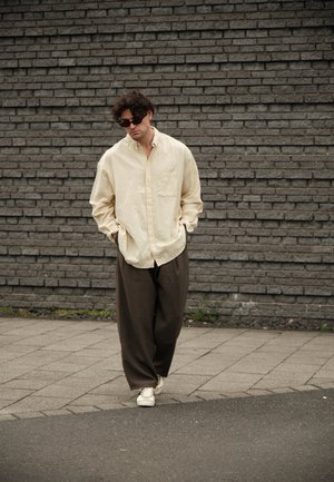 Hombre con camisa crema extragrande, pantalones marrones sueltos, zapatillas blancas y gafas de sol negras caminando por una acera contra una pared de ladrillo gris.