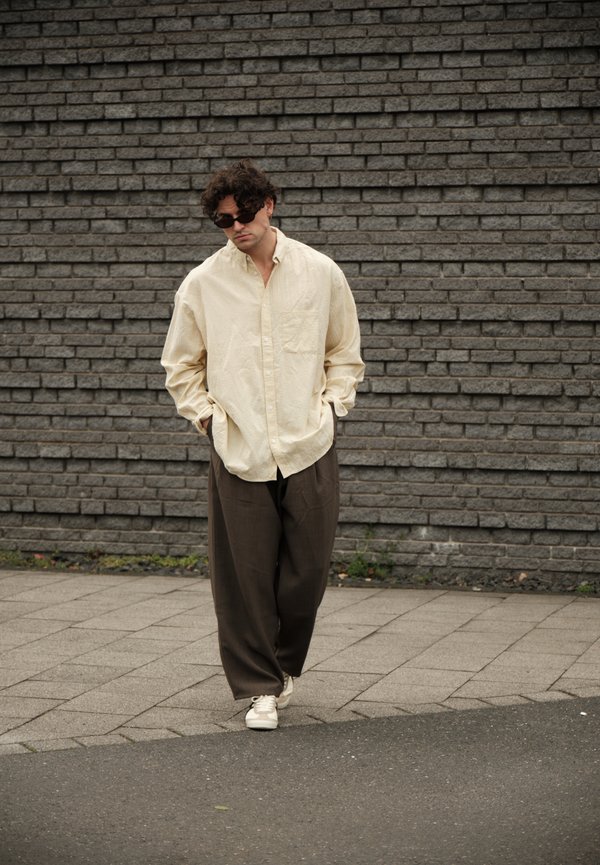 Férfi túlságosan nagy krémszínű ingben, bő barna nadrágban, fehér sportcipőben és fekete napszemüvegben sétál a járdán, egy szürke téglafal mellett.