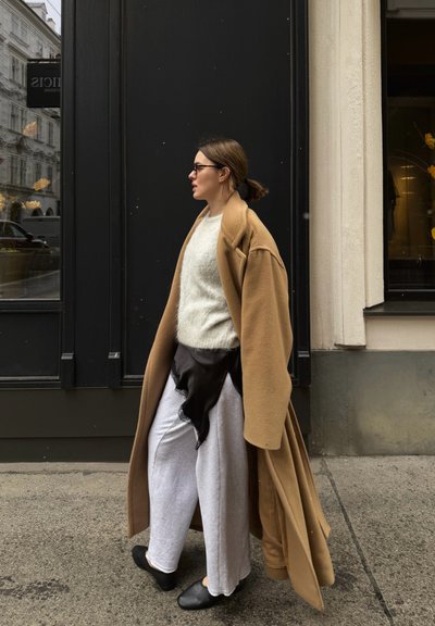 Mujer con cabello castaño recogido en un moño camina por la acera de la ciudad, lleva gafas, un abrigo largo color camel, suéter blanco, pantalones de varias capas y zapatos negros.