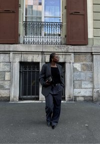Blazer noir surdimensionné par-dessus un haut noir ajusté, associé à un pantalon sombre ample et des escarpins noirs pointus ; portant une pochette noire.