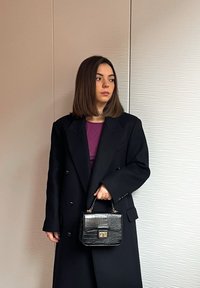 Femme aux cheveux bruns mi-longs portant un manteau long noir par-dessus un haut violet, tenant un petit sac à main en cuir noir, debout devant un mur texturé.